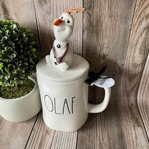 May include: White ceramic mug with a black "Olaf" inscription. The mug has a lid with a ceramic Olaf figurine on top. The mug has a black ribbon with a white "Frozen" tag attached.