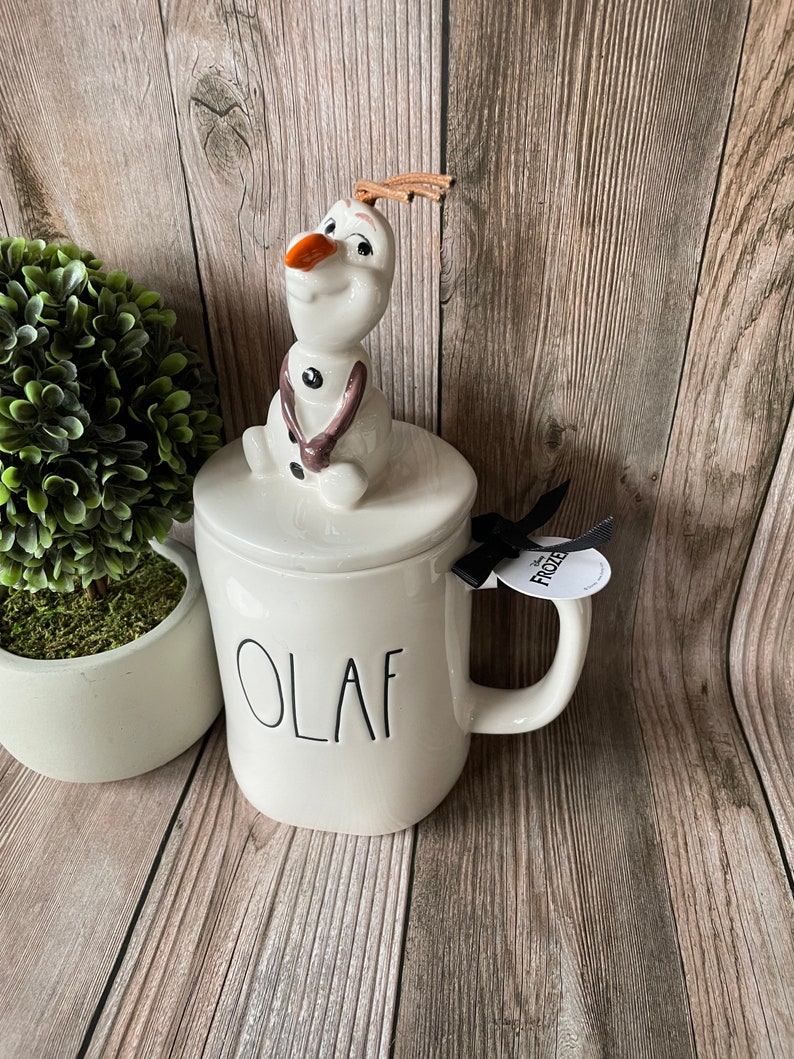 May include: White ceramic mug with a black "Olaf" inscription. The mug has a lid with a ceramic Olaf figurine on top. The mug has a black ribbon with a white "Frozen" tag attached.