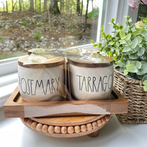 May include: Four white ceramic canisters with wooden lids and black lettering. The canisters are labeled "Rosemary" and "Tarragon". The canisters are sitting on a wooden tray with a beaded base.