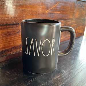 May include: A black ceramic mug with a curved handle. The word "SAVOR" is written in white, cursive lettering on the front of the mug. The mug is sitting on a dark wooden surface.
