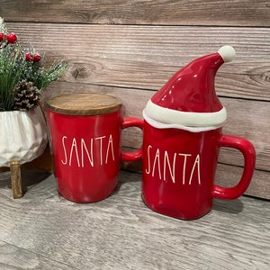 Puede incluir: Dos tazas de cer&aacute;mica roja con la inscripci&oacute;n "SANTA" en blanco. Una taza tiene una tapa de gorro de Pap&aacute; Noel rojo con ribete blanco.