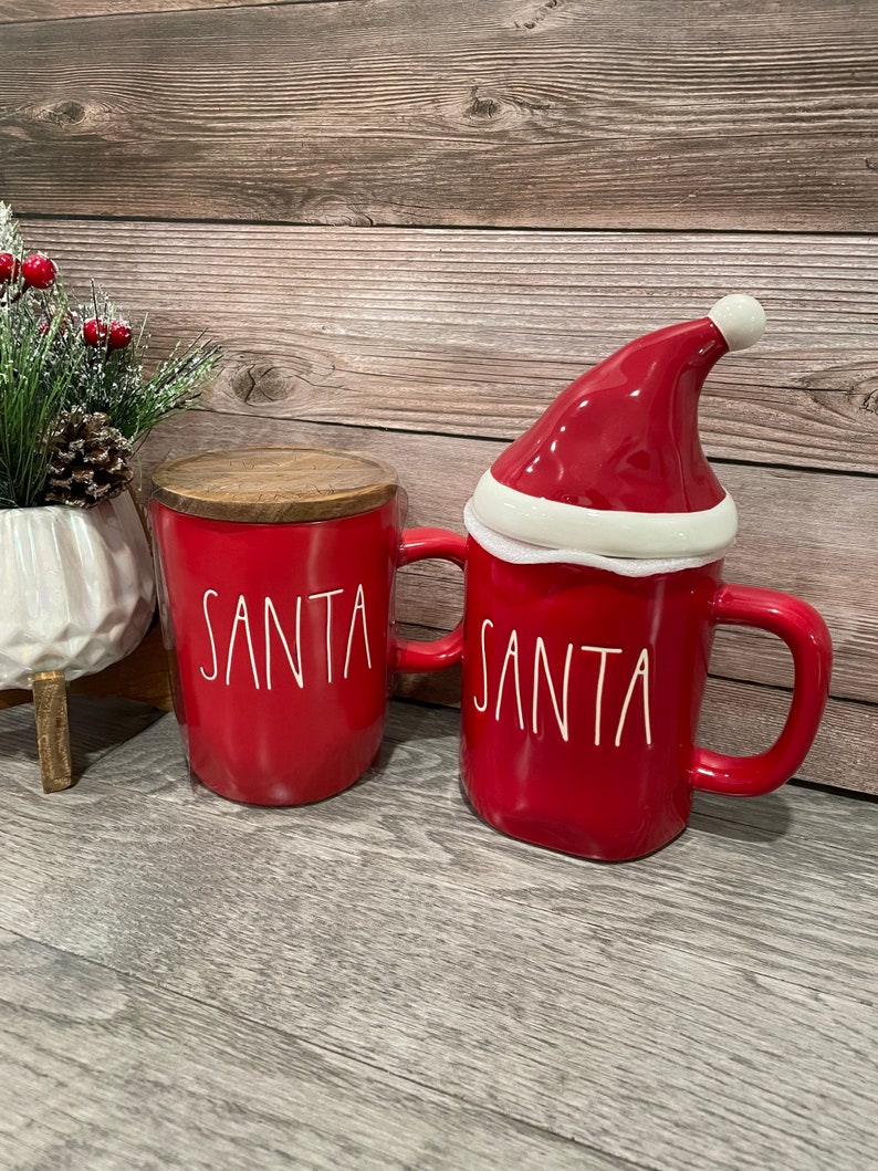 Puede incluir: Dos tazas de cer&aacute;mica roja con la inscripci&oacute;n "SANTA" en blanco. Una taza tiene una tapa de gorro de Pap&aacute; Noel rojo con ribete blanco.