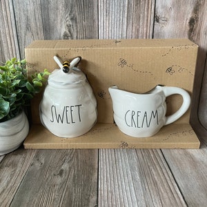 May include: A white ceramic sugar and creamer set with a bee design. The sugar container has a bee on top and says "SWEET" on the front. The creamer says "CREAM" on the front. The set is packaged in a brown box with a bee design.