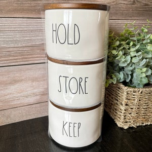 May include: A set of three white ceramic canisters with wooden lids. The canisters are stacked on top of each other. The top canister says "HOLD", the middle canister says "STORE", and the bottom canister says "KEEP".