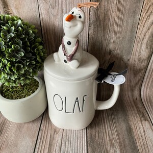 May include: White ceramic mug with a black "OLAF" inscription. The mug has a lid with a 3D Olaf figurine on top. The figurine is white with brown accents and has a carrot nose and twig arms.