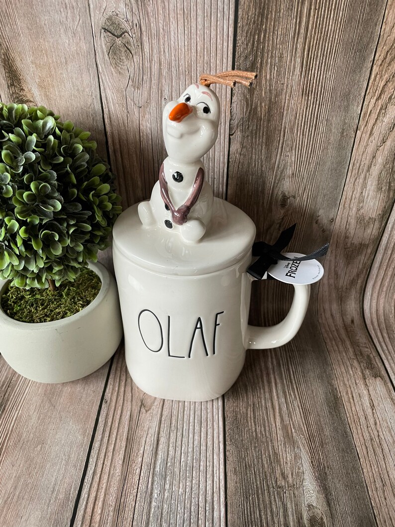 May include: White ceramic mug with a black "OLAF" inscription. The mug has a lid with a 3D Olaf figurine on top. The figurine is white with brown accents and has a carrot nose and twig arms.