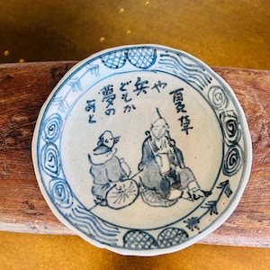 May include: A small, round, off-white ceramic plate with blue hand-painted illustrations and Japanese text. The plate features a scene with two figures and decorative border patterns. The plate is displayed on a wooden surface.
