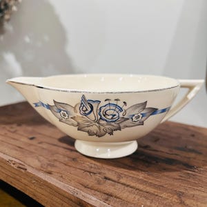 May include: Cream-colored gravy boat with a floral design in blue and gray. The boat has a spout, handle, and a pedestal base. A blue ribbon-like design encircles the boat, adding a decorative touch. The rim is trimmed with a silver-colored line.