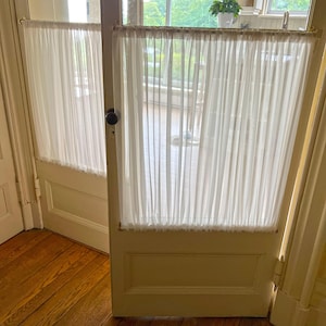 May include: Interior shot of a doorway with a sheer white curtain. The door is painted a light cream color and has a dark doorknob. The floor is made of wooden planks. The curtain is hung on a rod.