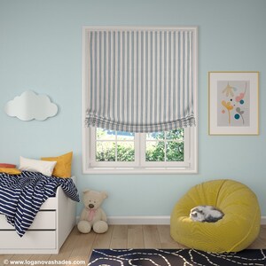 May include: A blue and white striped roman shade hangs in a window in a light blue room. The room has a yellow beanbag chair, a white bed with a blue and white striped comforter, a stuffed teddy bear, and a cloud-shaped wall decoration.