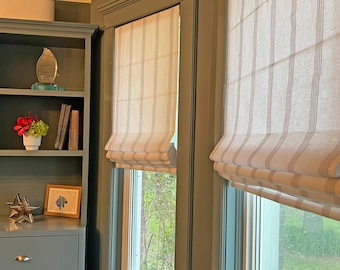 May include: Two off-white Roman shades featuring thin, vertical brown stripes installed on windows with dark green trim. The shades are partially raised, creating horizontal folds at the bottom, and are positioned next to a matching dark green cabinet.