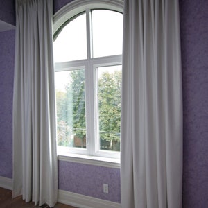May include: White, pleated curtains frame a large arched window. The curtains hang from a metal rod and are set against a purple textured wall. The window reveals a view of trees and a bright sky.