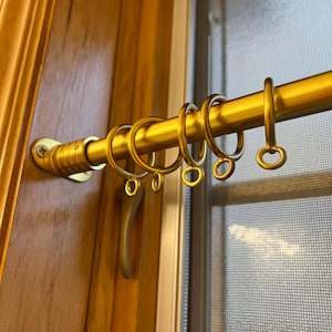 May include: A gold-colored curtain rod with matching rings and mounting hardware. The rod is mounted on a wooden door frame next to a window with a screen. The rings are designed to hold curtains.
