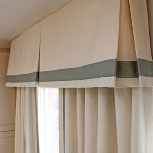 May include: Cream-colored valance with a gray border above matching curtains. The valance has a pleated design and is mounted above a window. The curtains are also cream-colored and hang in vertical folds.