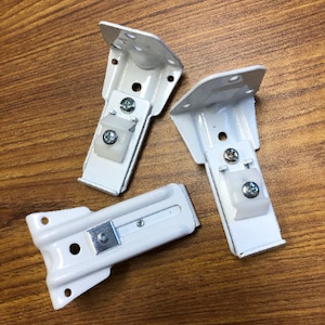 May include: Three white metal brackets with a screw and a white plastic piece on each. The brackets are designed to hold a curtain rod.