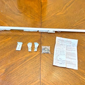 May include: A white curtain rod with an integrated pulley system and sliding gliders, accompanied by three metal mounting brackets, a small plastic bag containing screws, and a printed instruction sheet for installation and bracket usage.