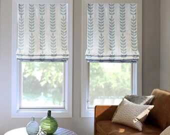 May include: Two white roman shade windows with a blue and white leaf pattern. The shades are pulled up to reveal a view of the outdoors. A brown leather sofa with pillows sits in front of the windows.