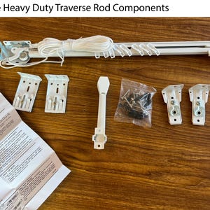 May include: A white heavy-duty traverse rod with a coiled white cord, four white metal mounting brackets, a white plastic baton, a small plastic bag containing metal screws and hardware, and an instruction manual labeled Single Heavy Duty Traverse Rod Components.