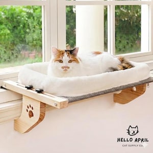 Pode incluir: Uma cama para gatos montada numa janela. A cama é branca e fofa, com uma base cinzenta. A estrutura de madeira tem um recorte de pegada. Um gato calico está a descansar na cama. O logótipo Hello April Cat Supply Co. está visível.