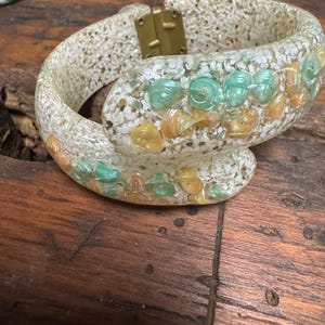May include: A white plastic bangle bracelet with a gold clasp. The bracelet is decorated with seashells and turquoise-colored stones.
