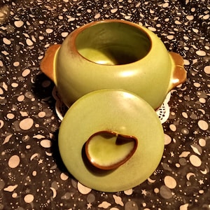 Puede incluir: Un plato de cerámica verde con tapa. El plato tiene un borde marrón y una perilla marrón en la tapa.