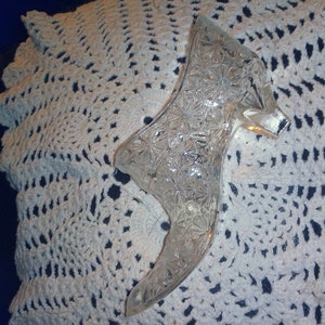 May include: A clear glass shoe-shaped dish with a cut-glass design. The dish is sitting on a white crocheted doily.