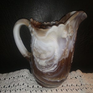 May include: A brown and white swirl glass pitcher with a scalloped rim and a floral design. The pitcher has a handle and is made of opaque glass.