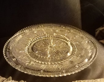 May include: A clear glass serving plate with a raised, intricate design. The plate has a scalloped edge and a central design with a starburst pattern.