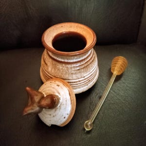 May include: A ceramic honey pot with a brown and white glaze. The pot has a lid with a bird design. A clear plastic honey dipper is included.