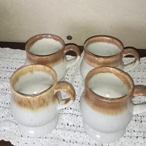 May include: Set of four ceramic mugs with a brown and white speckled glaze. The mugs have a rounded shape and a handle.