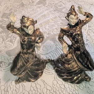 May include: Two ceramic figurines of people in traditional clothing. The figurines are painted in gold and black with white accents. They are posed in a dancing position.