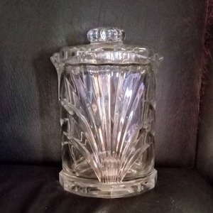 May include: A clear glass jar with a lid, featuring a decorative, geometric pattern. The jar has a wide base and a rounded top.