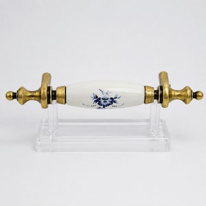 May include: A horizontal drawer pull featuring a central white ceramic handle with a blue floral pattern. The handle is flanked by two ornate, weathered brass-toned metal end pieces with rounded finials, connected by matching brass-toned hardware.