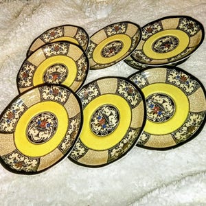 May include: Set of seven yellow and black patterned saucers with a gold rim. The saucers have a floral design with blue, red, and white accents.