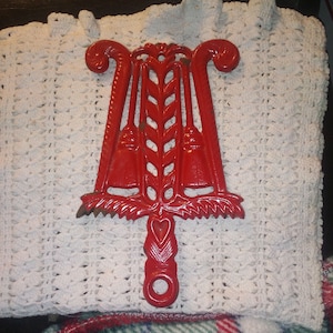 May include: Red cast iron trivet with a heart design and a wheat stalk pattern. The trivet has three loops on the top and a handle on the bottom.