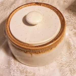 Puede incluir: Una cacerola de cerámica blanca con un borde marrón y una tapa. La tapa tiene un pequeño pomo en la parte superior.