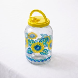 May include: A clear glass jar with a yellow lid and handle. The jar is decorated with yellow and blue sunflower designs. The base of the jar has a blue and white geometric pattern.
