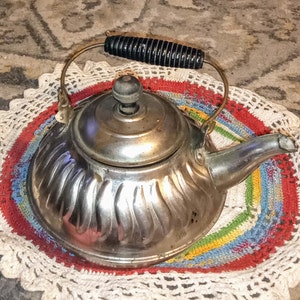 May include: A silver teapot with a black handle and a fluted design sits on a colorful crocheted doily.