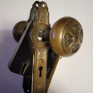 May include: Antique brass door knob set with a decorative floral design on the knob and backplate. The set includes two knobs and a backplate with a keyhole. The metal has a warm, aged patina, suggesting vintage craftsmanship.