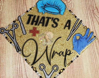 May include: A gold glitter graduation cap with the words "THAT'S A Wrap" in black script. The cap is decorated with a blue surgical cap, medical tools, a red cross, and a hand gesture.