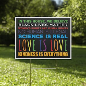Puede incluir: Un letrero rectangular para jard&iacute;n con fondo negro y texto multicolor que dice: IN THIS HOUSE, WE BELIEVE, BLACK LIVES MATTER, WOMEN'S RIGHTS ARE HUMAN RIGHTS, NO HUMAN IS ILLEGAL, SCIENCE IS REAL, LOVE IS LOVE, KINDNESS IS EVERYTHING.