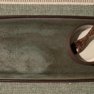 Puede incluir: Una bandeja de servicio rectangular de cerámica con una superficie esmaltada en verde oscuro y un borde marrón. Un pequeño cuenco redondo de cerámica blanca con el exterior marrón se sitúa a la derecha, conteniendo una cuchara de madera.