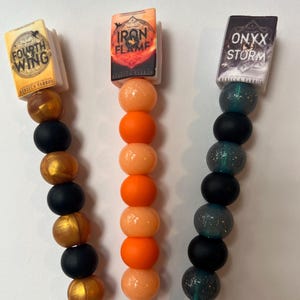 May include: Three crochet hooks with decorative handles featuring miniature book covers titled Fourth Wing, Iron Flame, and Onyx Storm by Rebecca Yarros. The handles are adorned with alternating spherical beads in metallic gold, matte black, solid orange, and glittery teal.