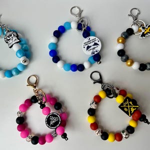 May include: A collection of five beaded wristlet keychains in various colors. Each keychain features a circular design with a clasp and a charm. The beads are in shades of blue, pink, black, gold, and yellow. Some charms have text and graphic designs.