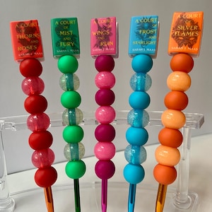 May include: Five colorful crochet hook toppers featuring miniature book covers with titles like "A Court of Thorns and Roses" and "A Court of Mist and Fury." Each hook has a different color scheme, including red, green, pink, blue, and orange beads.