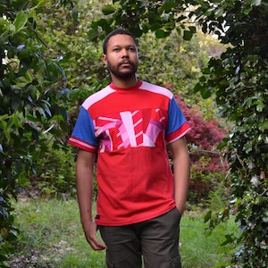 Puede incluir: Un hombre con una camisa de manga corta con estampado rojo, azul y rosa con cuello blanco. Está de pie en un entorno boscoso.