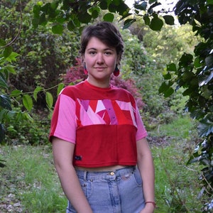 Könnte beinhalten: Eine Frau trägt ein rotes und pinkfarbenes, bauchfreies T-Shirt mit geometrischem Muster. Das Shirt hat kurze Ärmel und einen Rundhalsausschnitt. Sie trägt eine blaue Jeans und hat kurze braune Haare.