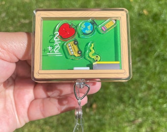 May include: A clear plastic badge holder with a green chalkboard design. The chalkboard has a red apple, a globe, a pencil, a yellow school bus, and a green worm. The badge holder has a silver retractable reel.