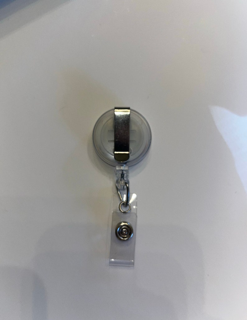 K&ouml;nnte beinhalten: Ein transparenter, einziehbarer Ausweishalter aus Kunststoff mit einem silbernen Clip und einem silbernen Knopf. Der Halter ist an einer silbernen Kette befestigt.