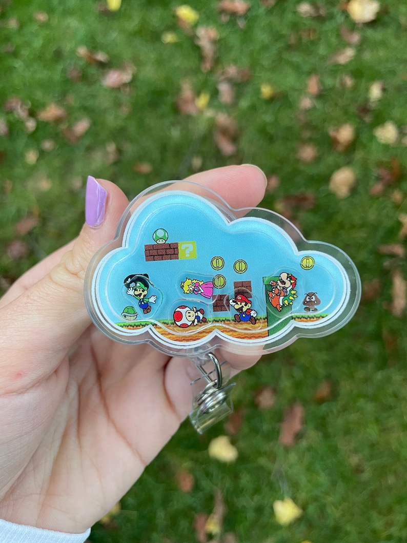 K&ouml;nnte beinhalten: Ein durchsichtiger Badgehalter aus Plastik mit einem blauen Wolken-Design, das Figuren aus dem Videospiel Super Mario Bros. zeigt. Die Wolke hat einen Fragezeichenblock, M&uuml;nzen und ein gr&uuml;nes Rohr. Die Figuren sind Mario, Luigi, Peach, Toad und Bowser.
